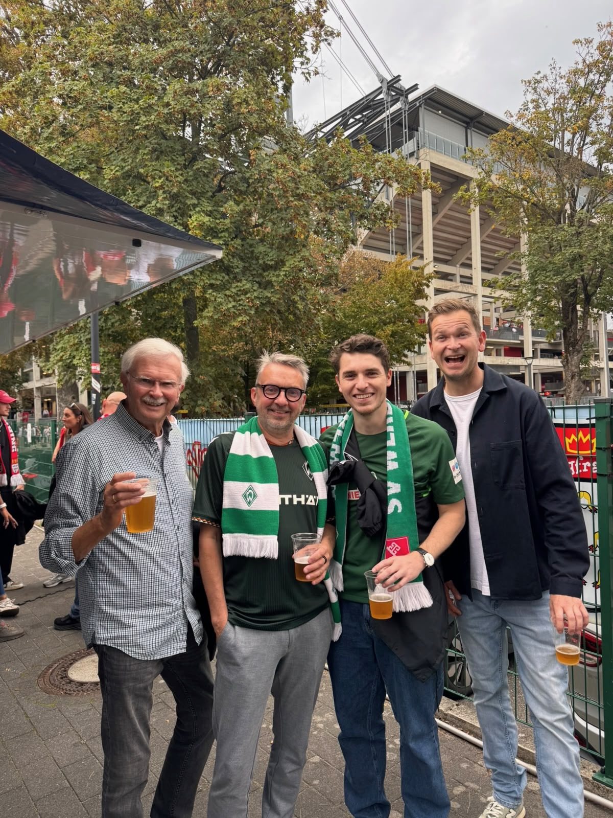 Vier Männer mit Bierbechern vor dem RheinEnergieStadion, zwei von ihnen in grün-weißen Werder-Trikots und -Schals.