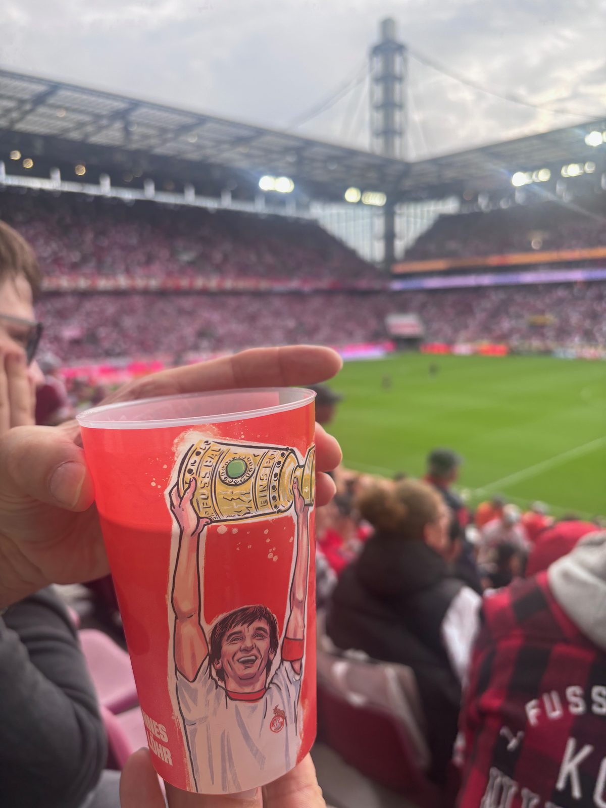 Ein roter Pfandbecher mit gezeichnetem Pokalsieger-Motiv wird vor den vollen Rängen des RheinEnergieStadions hochgehalten.