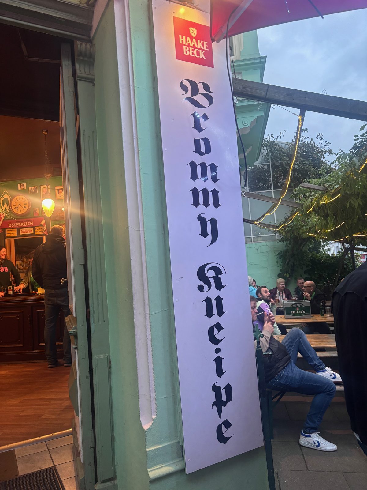 Hochkantes Schild mit der Aufschrift „Brommy Kneipe“ in gebrochener Schrift und Haake-Beck-Logo neben der offenen Tür der Kneipe, rechts Gäste an Biertischen unter einer Lichterkette.