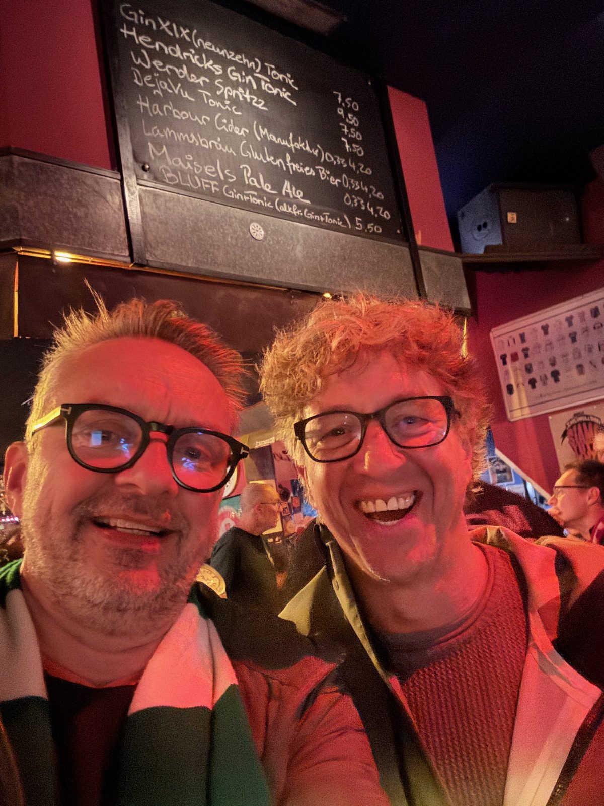 Selfie von zwei lachenden Männern in einer rot beleuchteten Kneipe, einer mit grün-weißem Werder-Schal. Über ihnen hängt eine Kreidetafel mit handgeschriebener Getränkekarte.