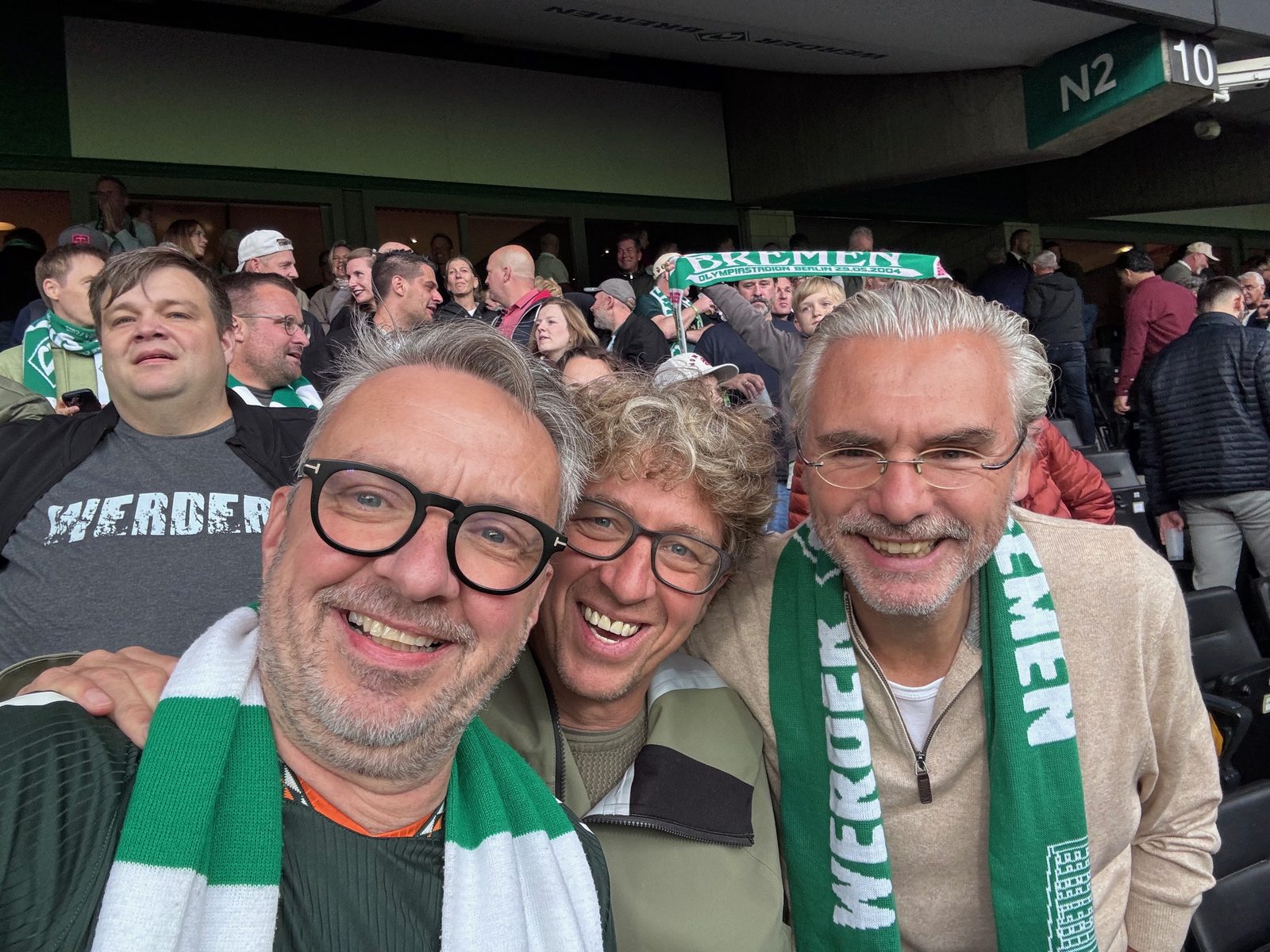 Selfie von drei lachenden Männern mit grün-weißen Werder-Schals auf der Tribüne des Weserstadions, hinter ihnen weitere Zuschauer und ein Schal mit der Aufschrift Bremen.