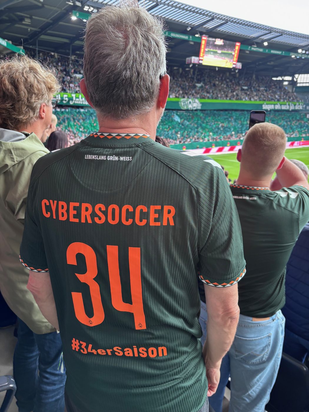 Rückenansicht eines dunkelgrünen Werder-Trikots mit der orangefarbenen Aufschrift CYBERSOCCER, der Nummer 34 und dem Schriftzug #34erSaison; darüber im Nacken „Lebenslang Grün-Weiss“, im Hintergrund die gefüllten Ränge und der Rasen des Weserstadions.