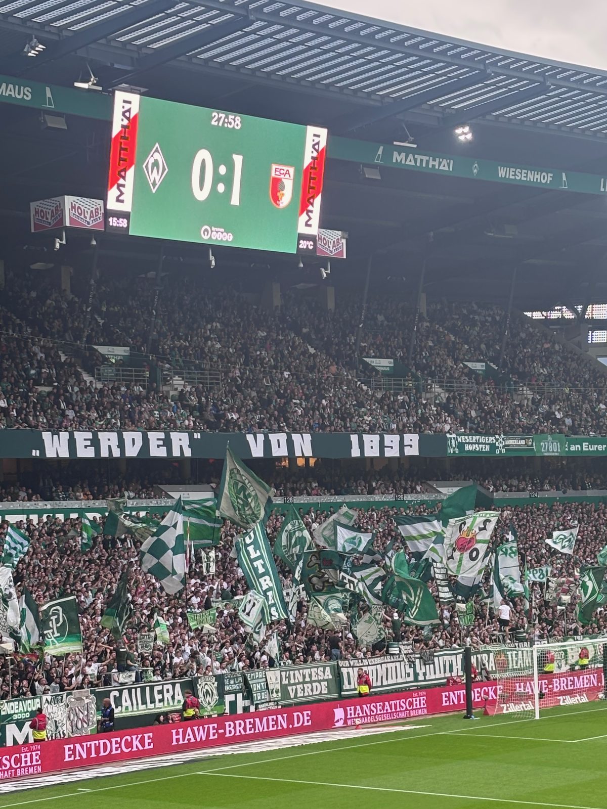 Die Anzeigetafel im Weserstadion zeigt den Stand 0:1 zwischen Werder Bremen und dem FC Augsburg. Darunter die Ostkurve mit dutzenden geschwenkten grün-weißen Fahnen und dem Banner „Werder von 1899“.