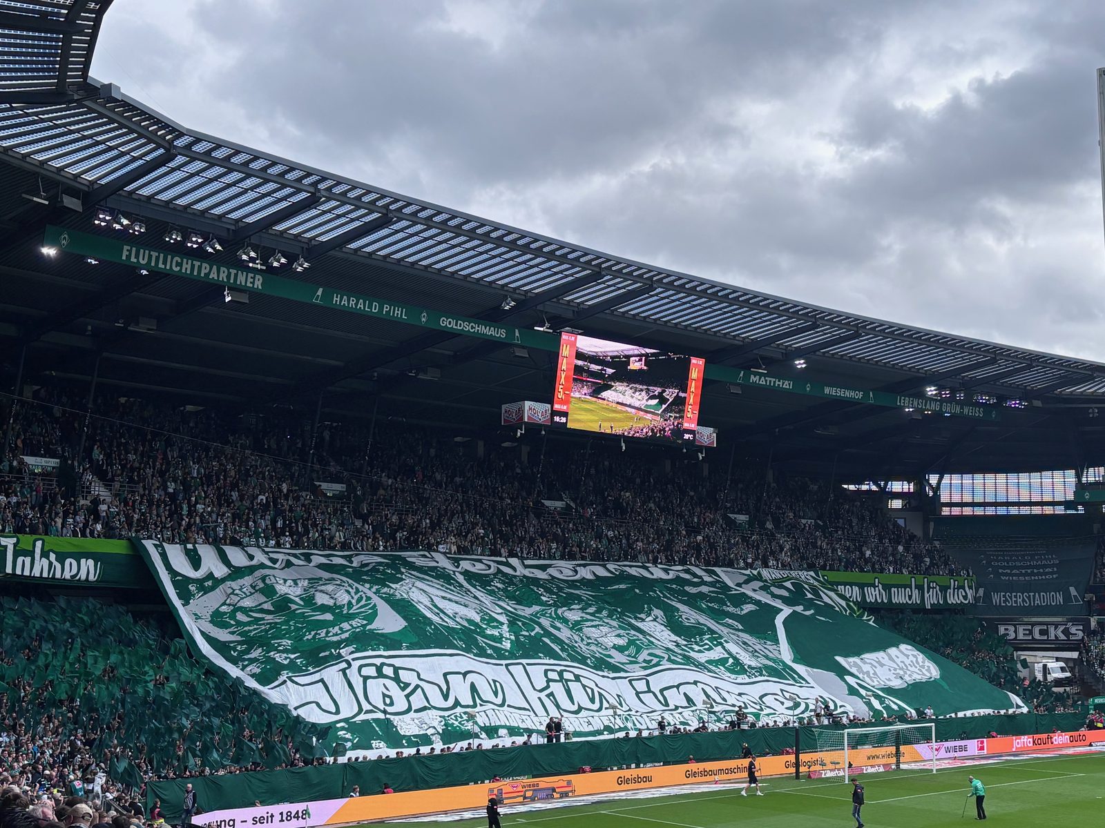 Die Ostkurve des Weserstadions ist vor dem Anpfiff vollständig von einer großen grün-weißen Blockfahne mit einem Porträt und einem Namenszug bedeckt, darüber ein Spruchband und die voll besetzten Ränge.