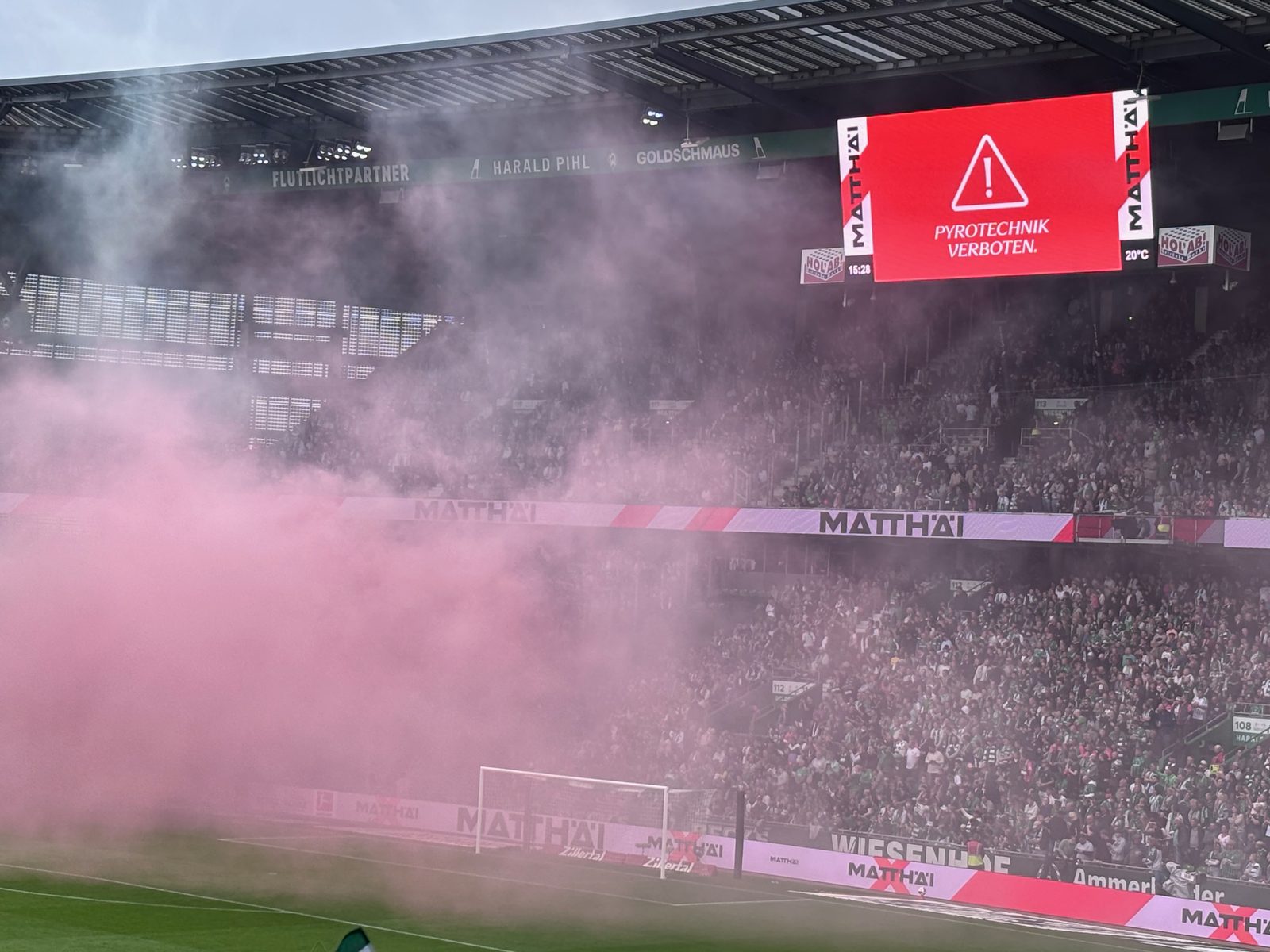 Dichter pinkfarbener Rauch zieht über den Rasen und die Tribüne des Weserstadions. Rechts oben leuchtet die Anzeigetafel rot mit einem Warndreieck und dem Text „Pyrotechnik verboten.“