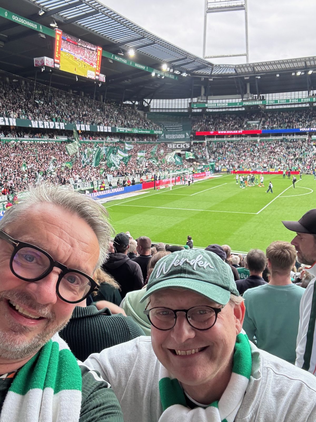 Selfie von zwei Männern mit grün-weißen Werder-Schals auf der Tribüne des Weserstadions, einer trägt eine grüne Werder-Kappe. Im Hintergrund die volle Ostkurve mit Fahnen und die Mannschaft auf dem Rasen.
