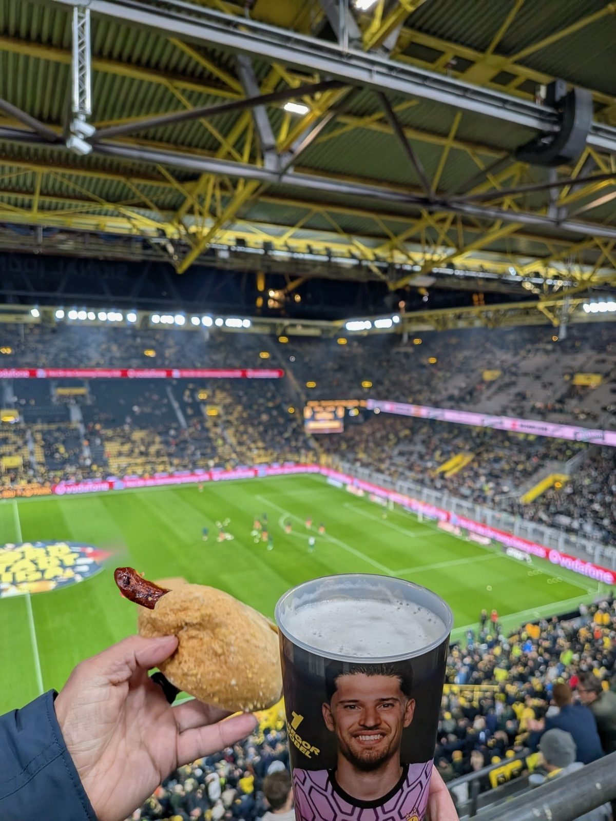 Eine Hand hält eine Wurst im Brötchen, daneben ein Bierbecher mit dem Bild von Torwart Gregor Kobel. Dahinter der Blick von oben auf Rasen und gelbe Ränge des Signal Iduna Parks.