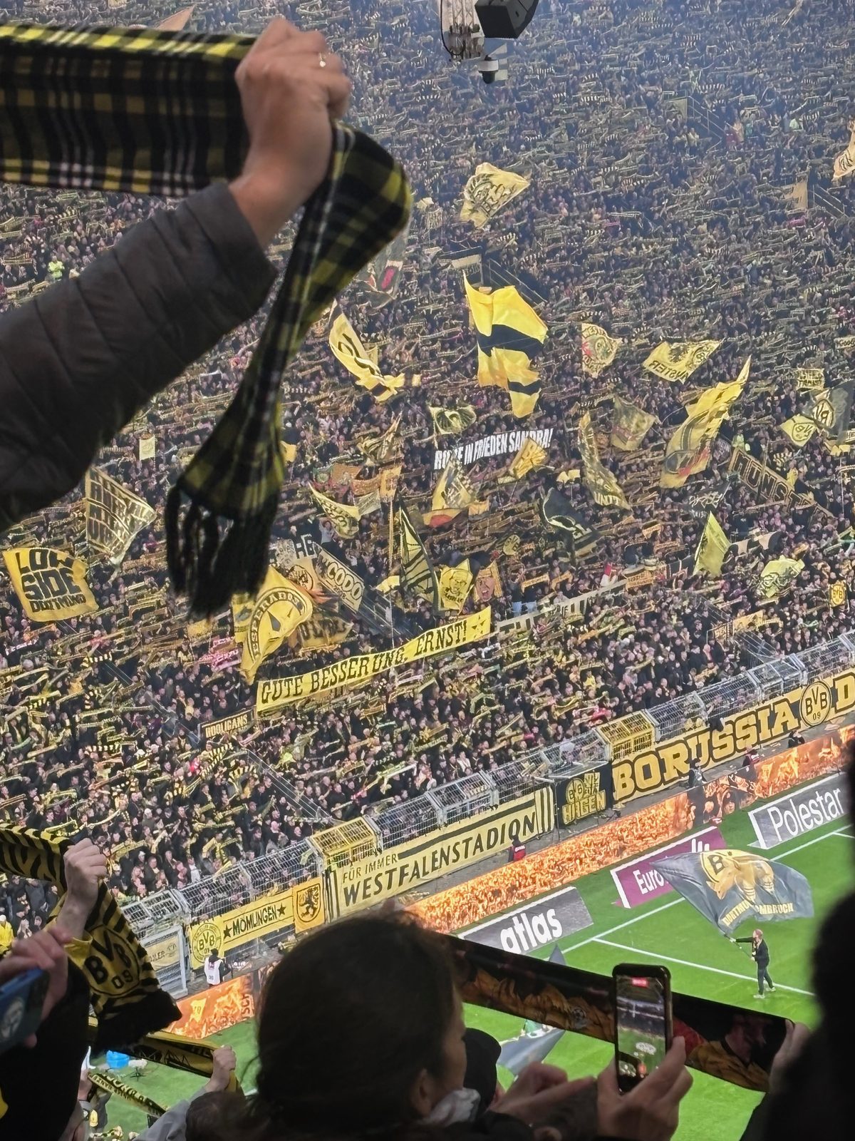 Die Südtribüne in Dortmund, dicht gefüllt mit schwarz-gelben Fahnen und Spruchbändern. Im Vordergrund recken Zuschauer Schals in die Höhe und filmen mit dem Handy.