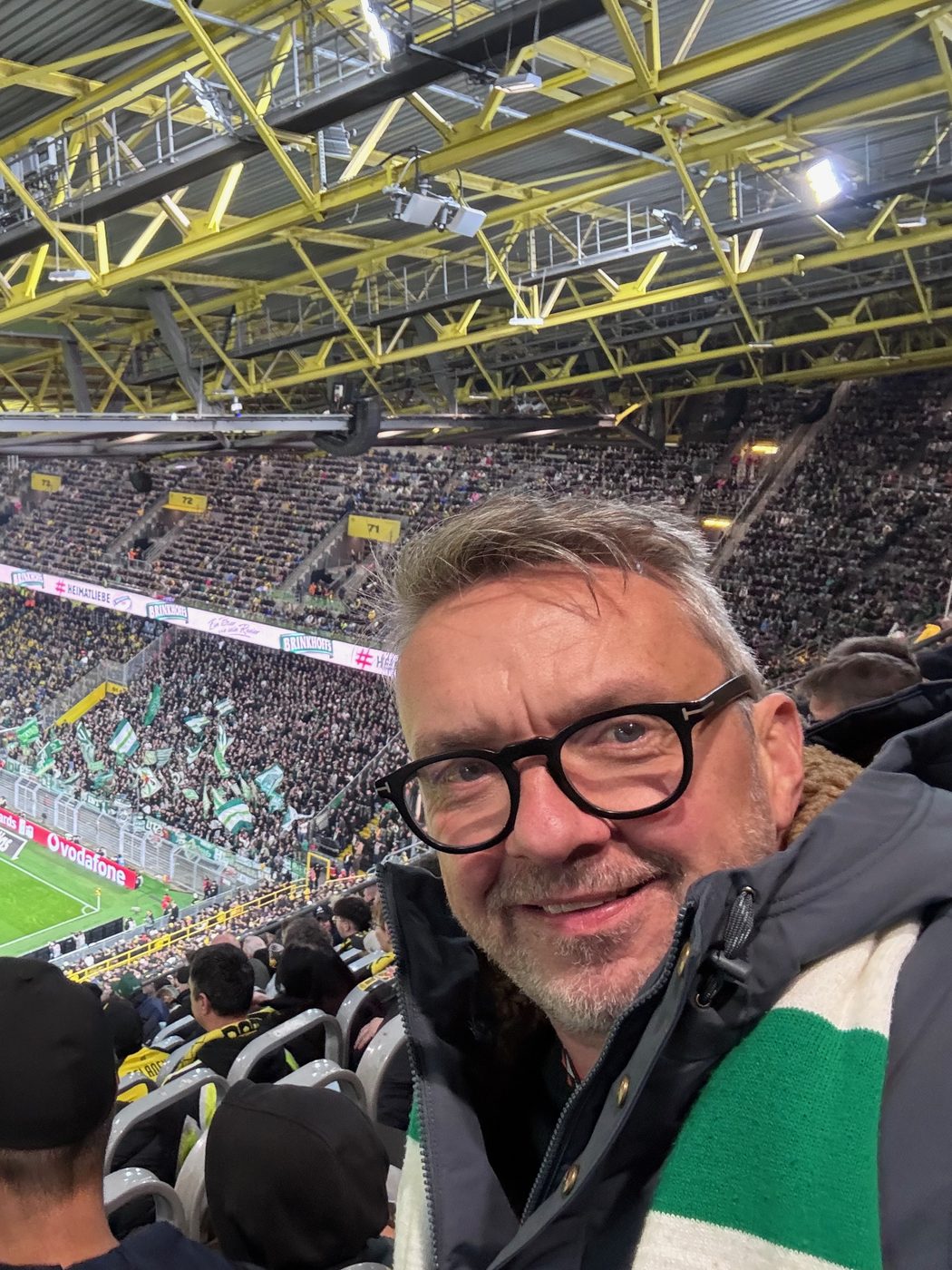 Selfie von Matthias mit Brille und grün-weißem Werder-Schal auf der Tribüne des Signal Iduna Parks. Hinter ihm die gelben Dachträger und der voll besetzte Gästeblock mit grün-weißen Fahnen, unten links ein Stück Rasen.