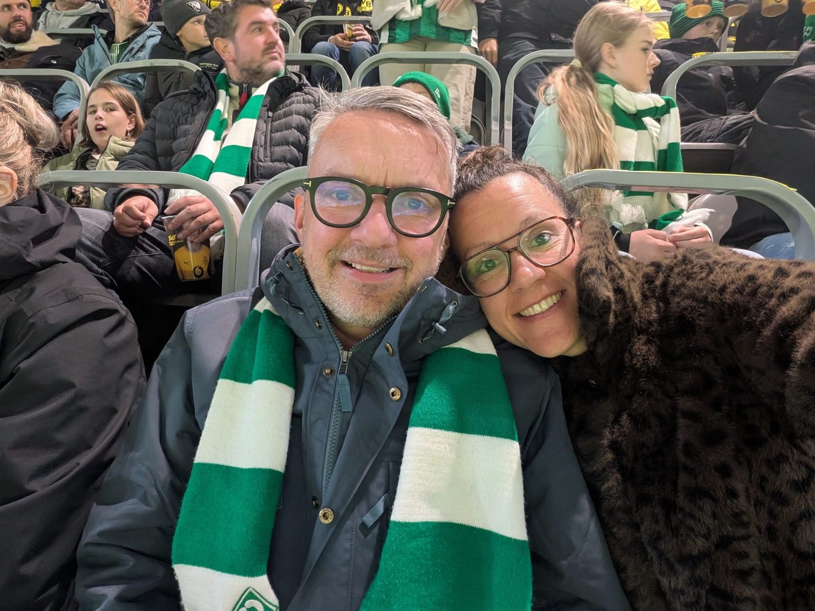 Selfie von Matthias mit grün-weißem Werder-Schal und Lena, die ihren Kopf an seine Schulter lehnt, beide lächelnd auf der Tribüne. Um sie herum weitere Zuschauer mit Werder-Schals.