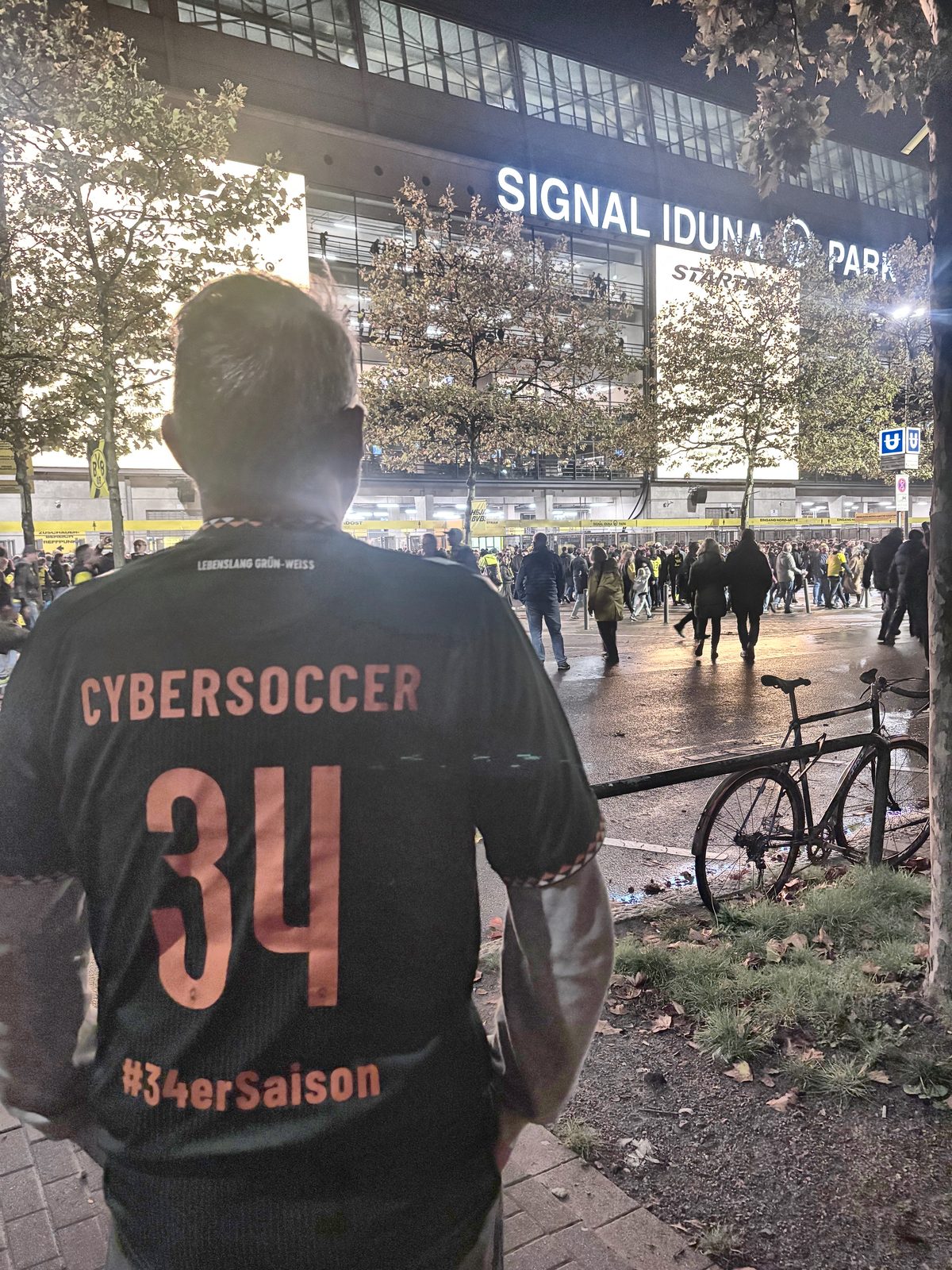 Rückenansicht eines dunklen Werder-Trikots mit der orangefarbenen Aufschrift CYBERSOCCER, der Nummer 34 und dem Schriftzug #34erSaison. Im Hintergrund der beleuchtete Schriftzug SIGNAL IDUNA PARK über dem Stadioneingang und Fans auf dem Vorplatz.