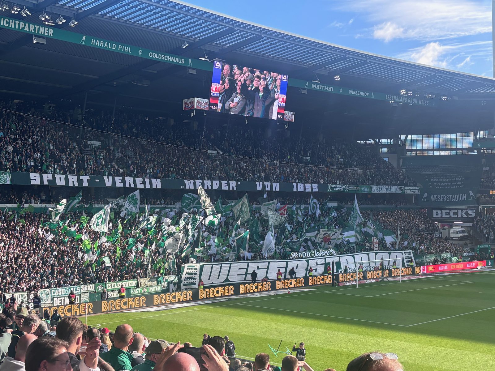 Die Ostkurve des Weserstadions vor dem Anpfiff: Der Unterrang ist voller grün-weißer Fahnen, davor ein großes Spruchband mit dem Schriftzug WESERSTADION, darüber das Band „Sport-Verein Werder von 1899“ und die Anzeigetafel.