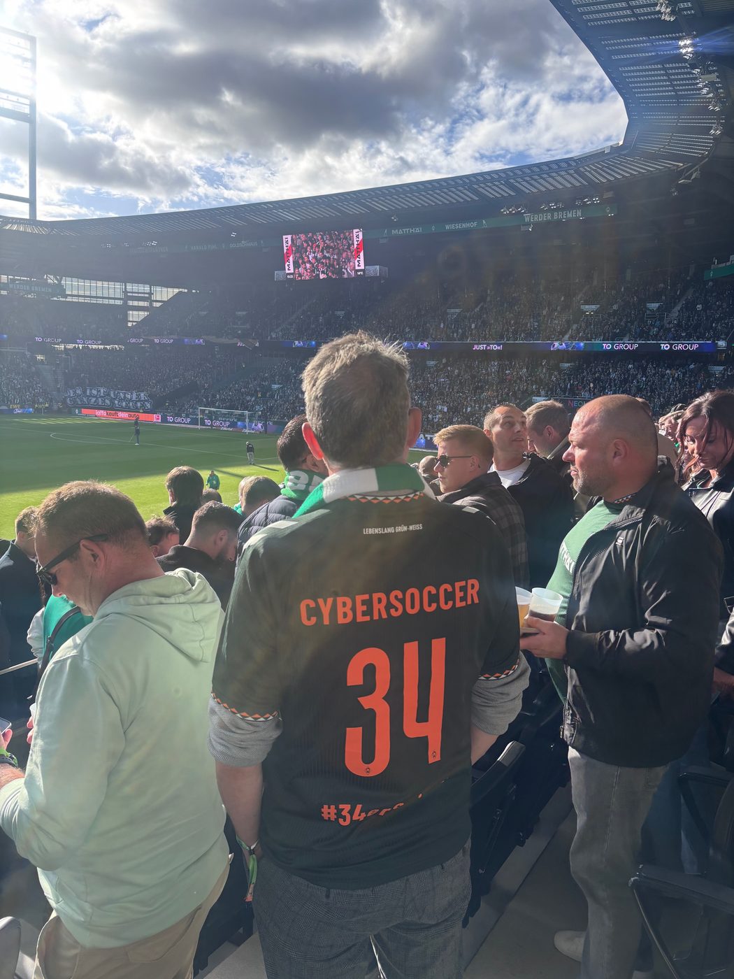 Rückenansicht von Matthias im dunklen Werder-Trikot mit der orangefarbenen Aufschrift CYBERSOCCER und der Nummer 34, grün-weißer Schal um den Hals. Er steht zwischen anderen Zuschauern auf der Tribüne des Weserstadions, dahinter Rasen, die volle Gegentribüne und die Anzeigetafel unter wolkigem Himmel.