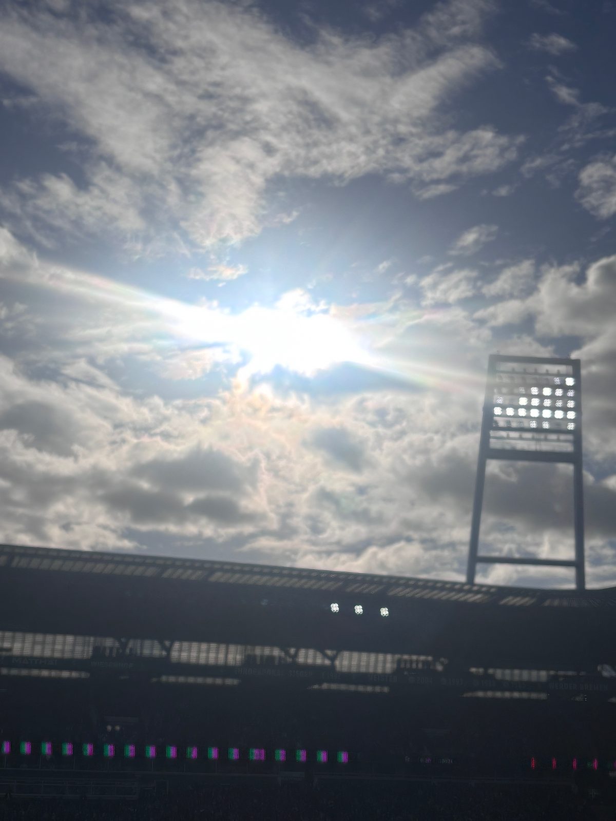 Gegenlichtaufnahme über dem Tribünendach des Weserstadions: Die Sonne bricht durch lockere Wolken, rechts ragt ein Flutlichtmast mit leuchtenden Scheinwerfern in den blauen Himmel.