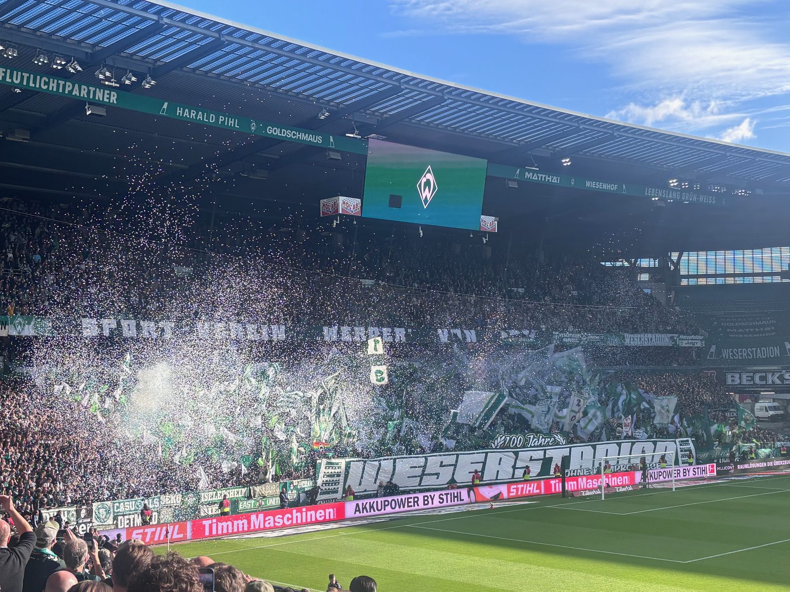 Die Ostkurve mit den grün-weißen Fahnen und dem WESERSTADION-Spruchband, über den Fahnen steigen erste weiße Rauchschwaden und Konfetti auf. Auf der Anzeigetafel das Werder-Wappen.