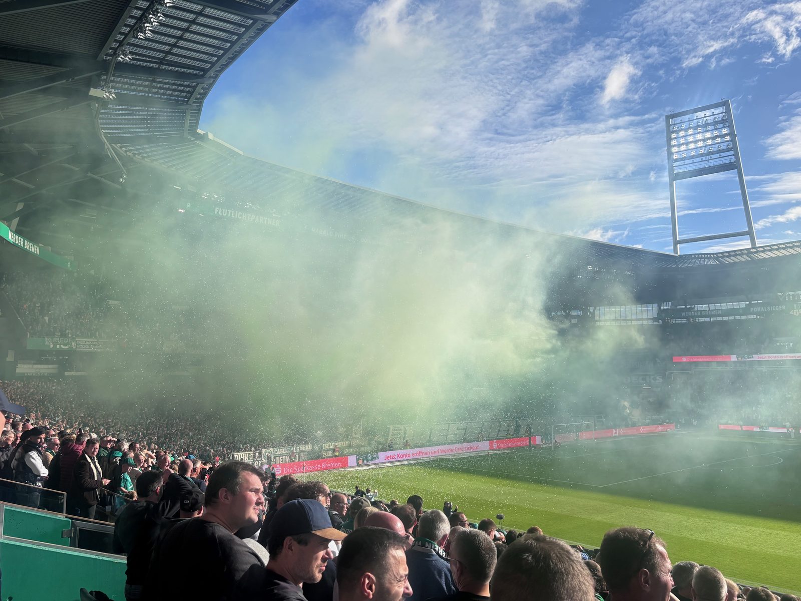 Grünlich-weißer Rauch hat die Ostkurve fast vollständig verhüllt und zieht über den Rasen, rechts der Flutlichtmast vor blauem Himmel, im Vordergrund Zuschauer auf der Tribüne.
