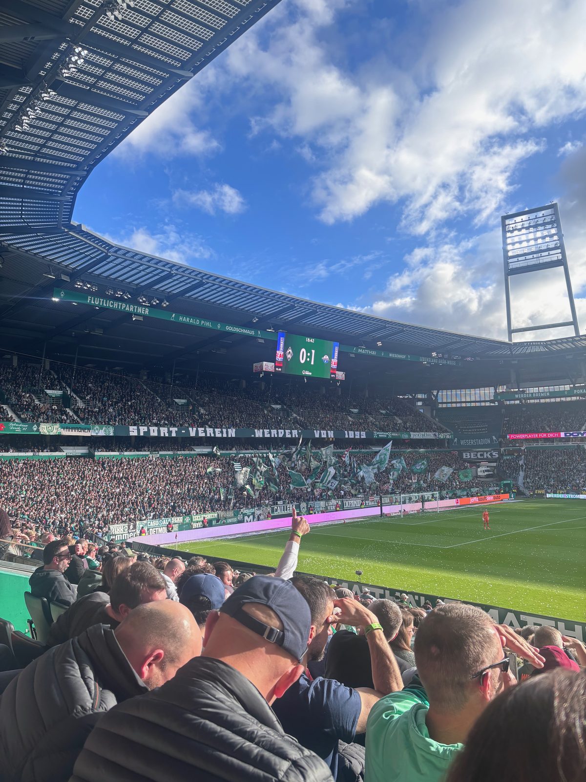 Blick von der Tribüne über den sonnigen Rasen auf die Ostkurve, die Anzeigetafel zeigt 0:1. Im Vordergrund Zuschauer, einer hebt den Arm.