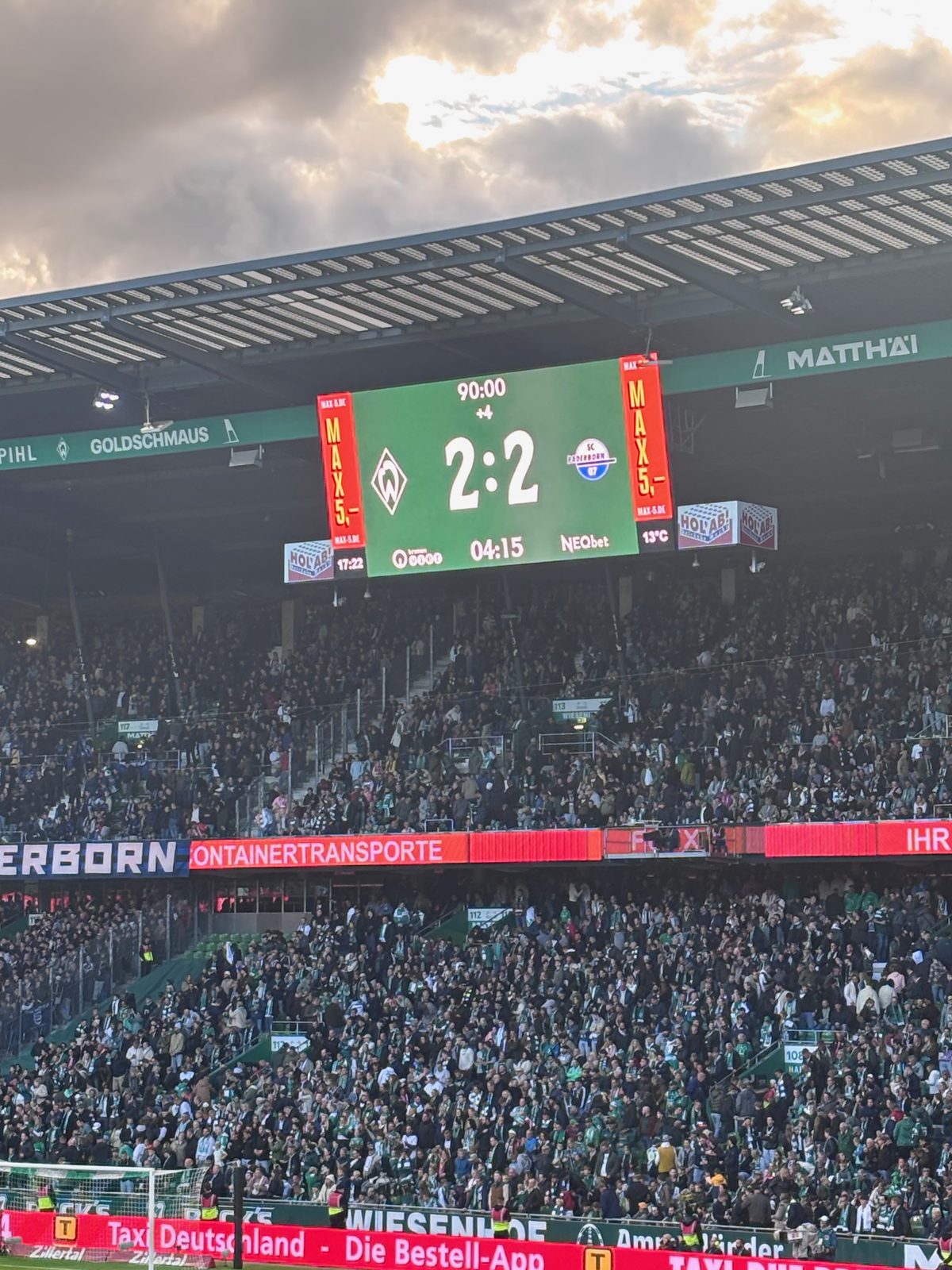 Nahaufnahme der Anzeigetafel über der vollen Ostkurve: Werder-Wappen, 2:2, Paderborn-Wappen, darüber Abendwolken.