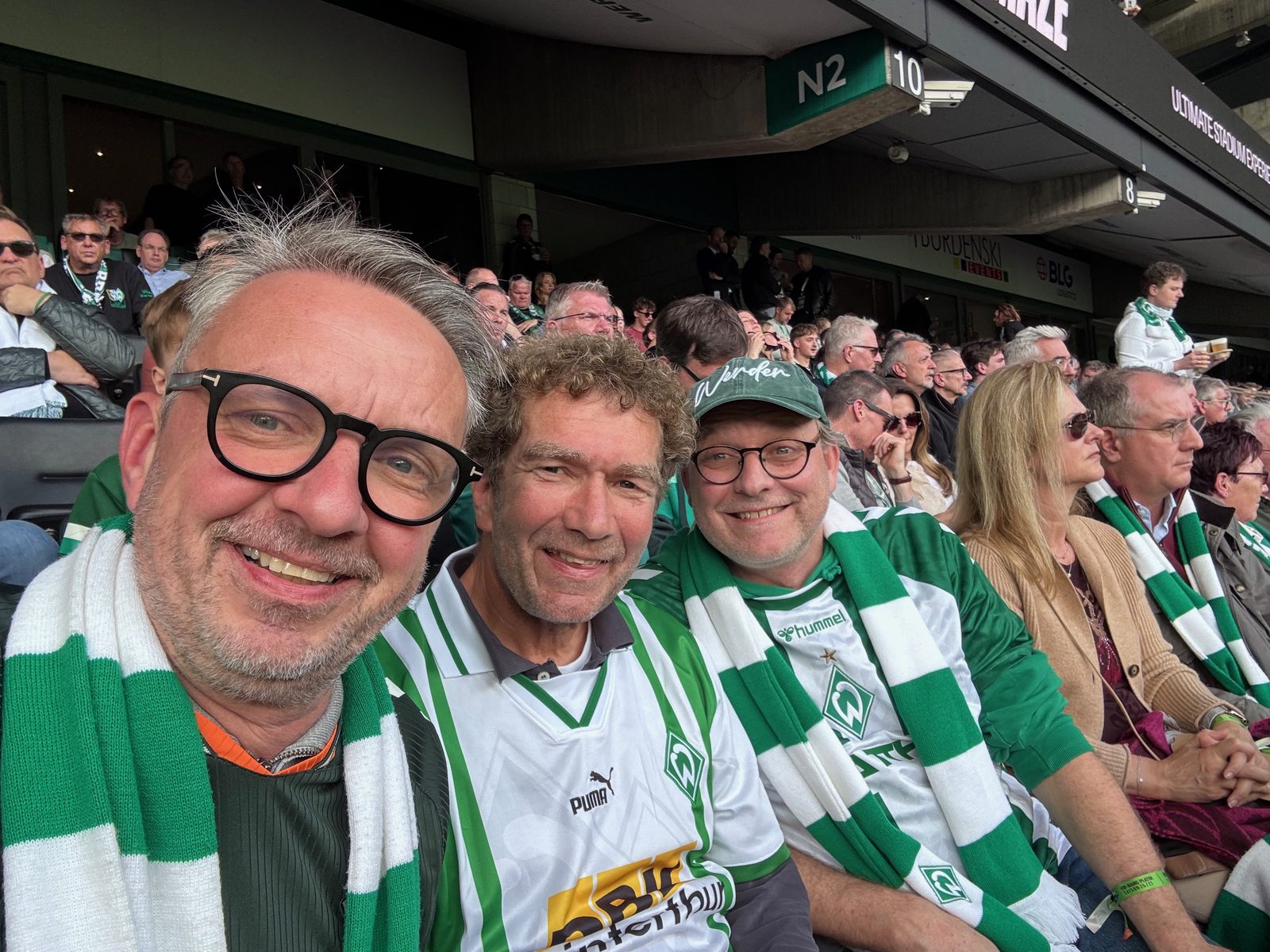 Selfie von drei Männern mit grün-weißen Werder-Schals auf der Tribüne des Weserstadions: links Matthias mit schwarzer Brille, in der Mitte Axel mit lockigen Haaren im weißen Werder-Trikot, rechts Thomas mit grüner Werder-Kappe. Dahinter weitere Zuschauer und das Blockschild N2.