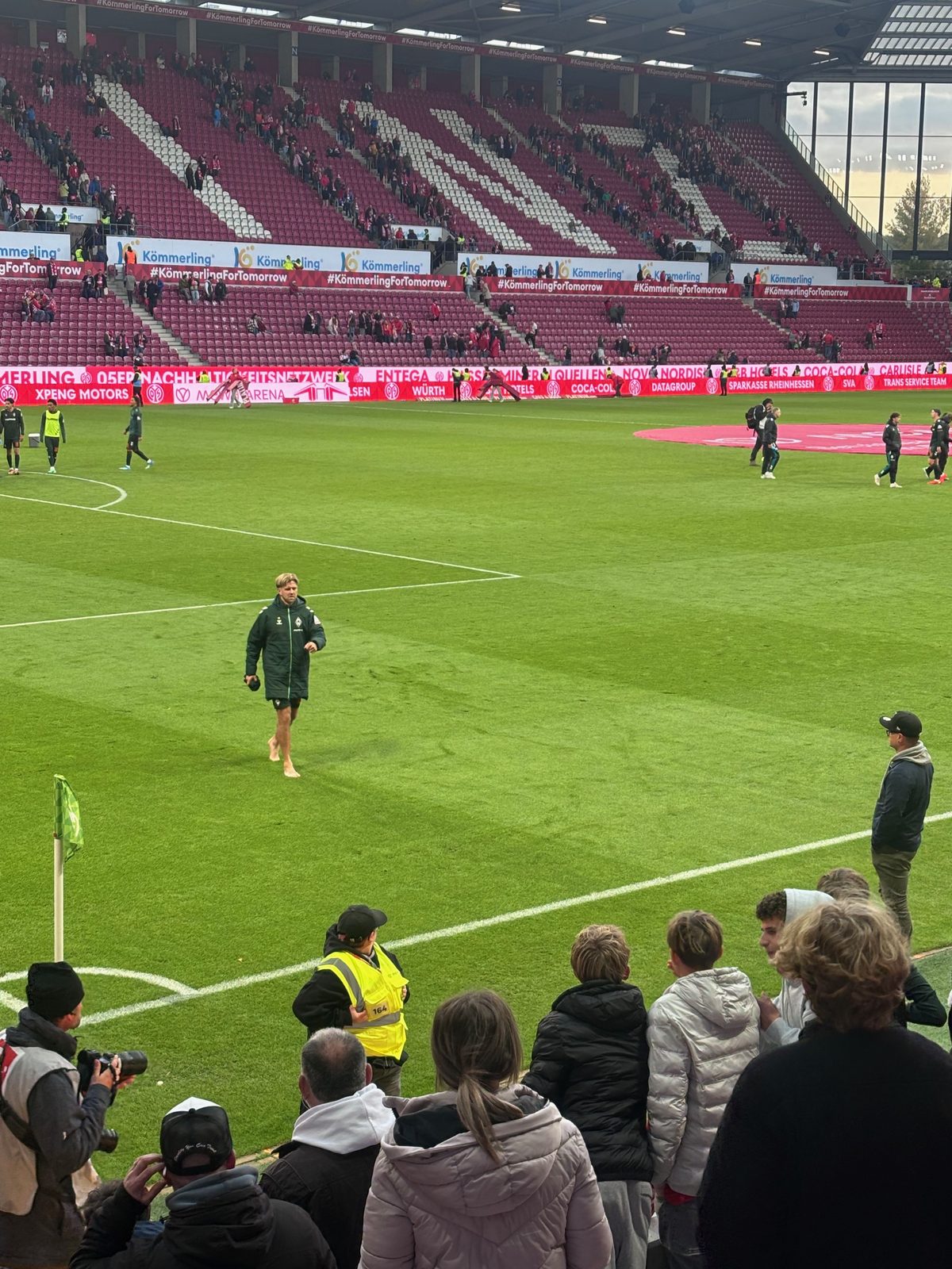 Nach dem Abpfiff: Niclas Füllkrug in grüner Trainingsjacke geht barfuß über den Rasen in Richtung Eckfahne und Tribüne, im Vordergrund Zuschauer, ein Fotograf und ein Ordner, dahinter die sich leerende Gegentribüne.