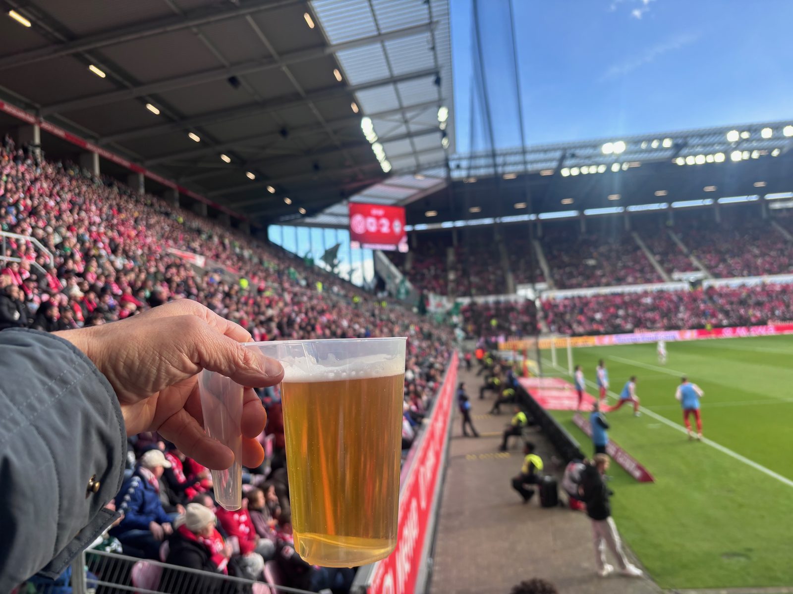 Eine Hand hält einen vollen Bierbecher vor die Tribüne, im Hintergrund zeigt die Anzeigetafel 0:2, rechts der Rasen und Spieler beim Aufwärmen an der Seitenlinie.