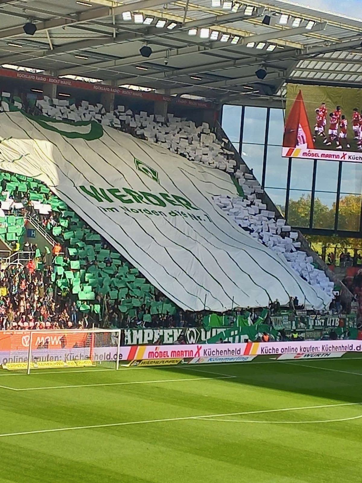Der Gästeblock frontal: Eine riesige weiße Blockfahne im Trikot-Design mit Werder-Raute und dem Schriftzug WERDER, ringsum grüne und weiße Papptafeln, darüber die Videowand.