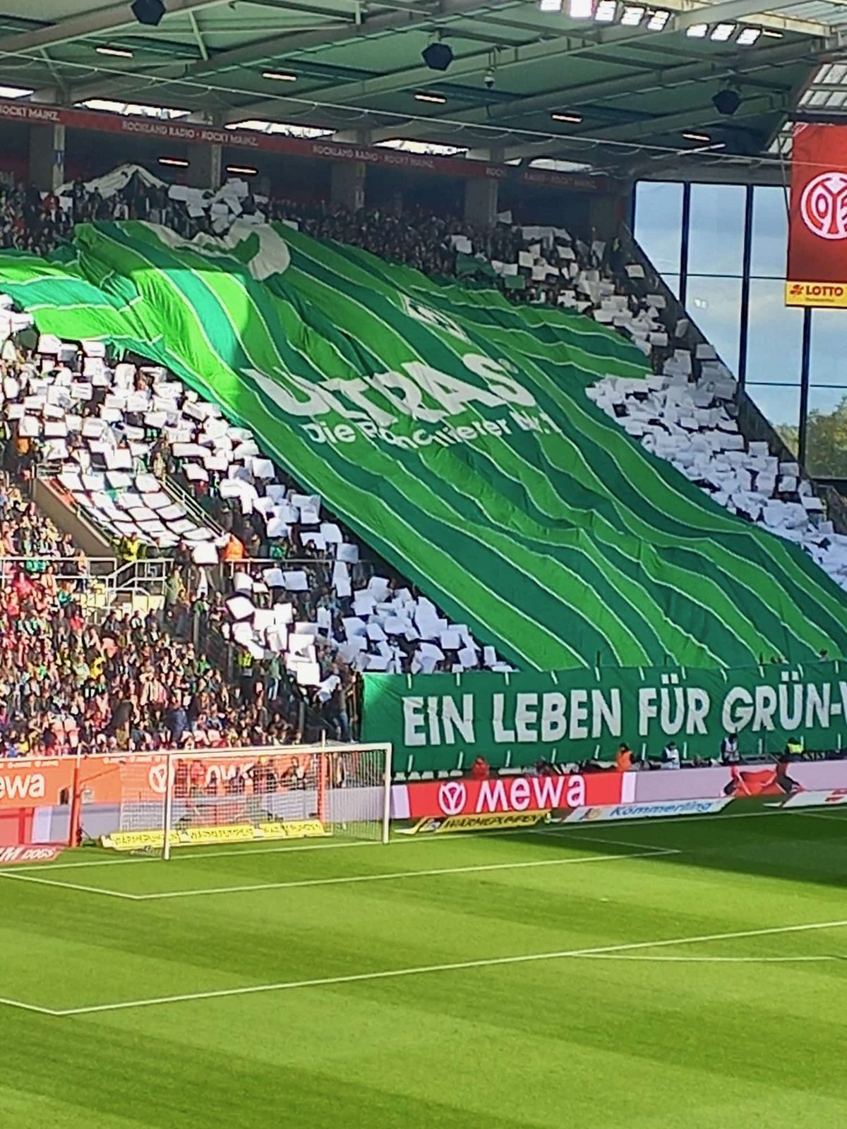 Der Gästeblock frontal: Eine riesige grün gestreifte Blockfahne, ringsum weiße Papptafeln, davor ein langes Spruchband „Ein Leben für Grün-Weiß“ über dem Tor.