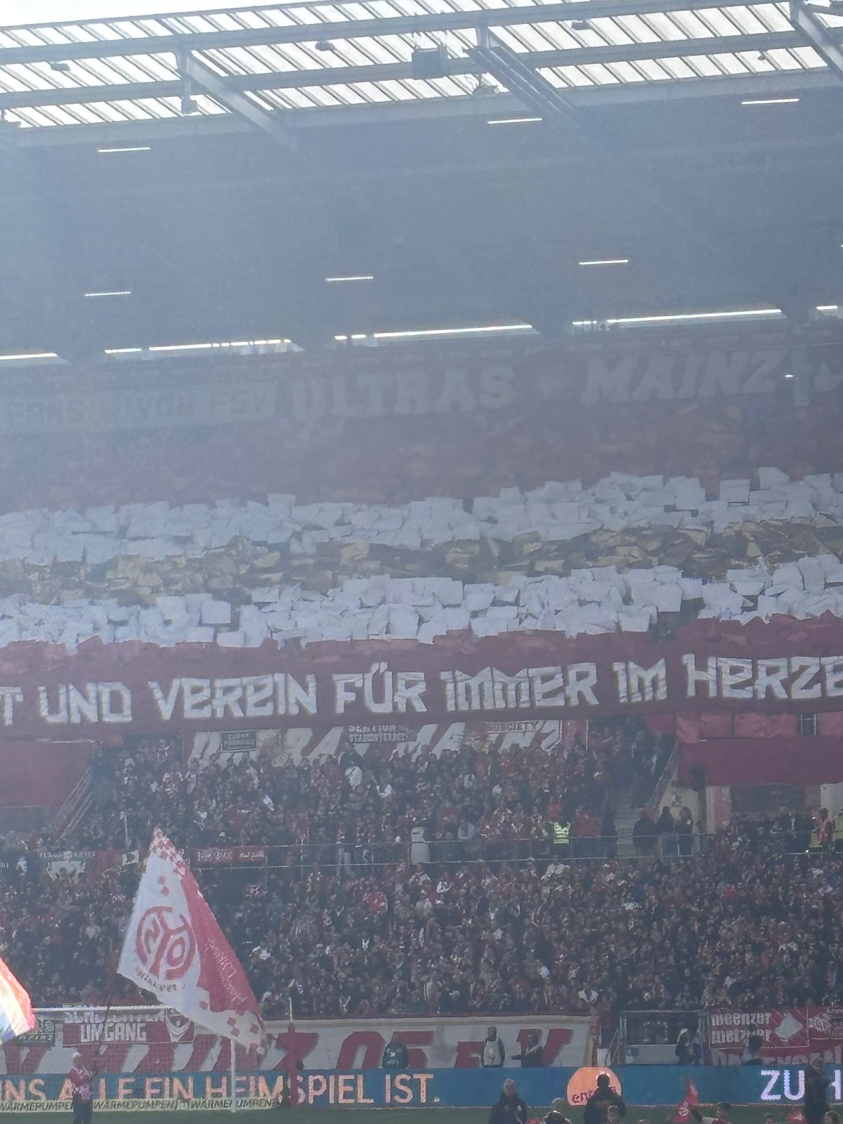 Die Mainzer Kurve im Gegenlicht: weiße und rote Papptafeln in Streifen, darunter ein rotes Spruchband „… und Verein für immer im Herzen“, vorne eine große Mainz-05-Fahne.