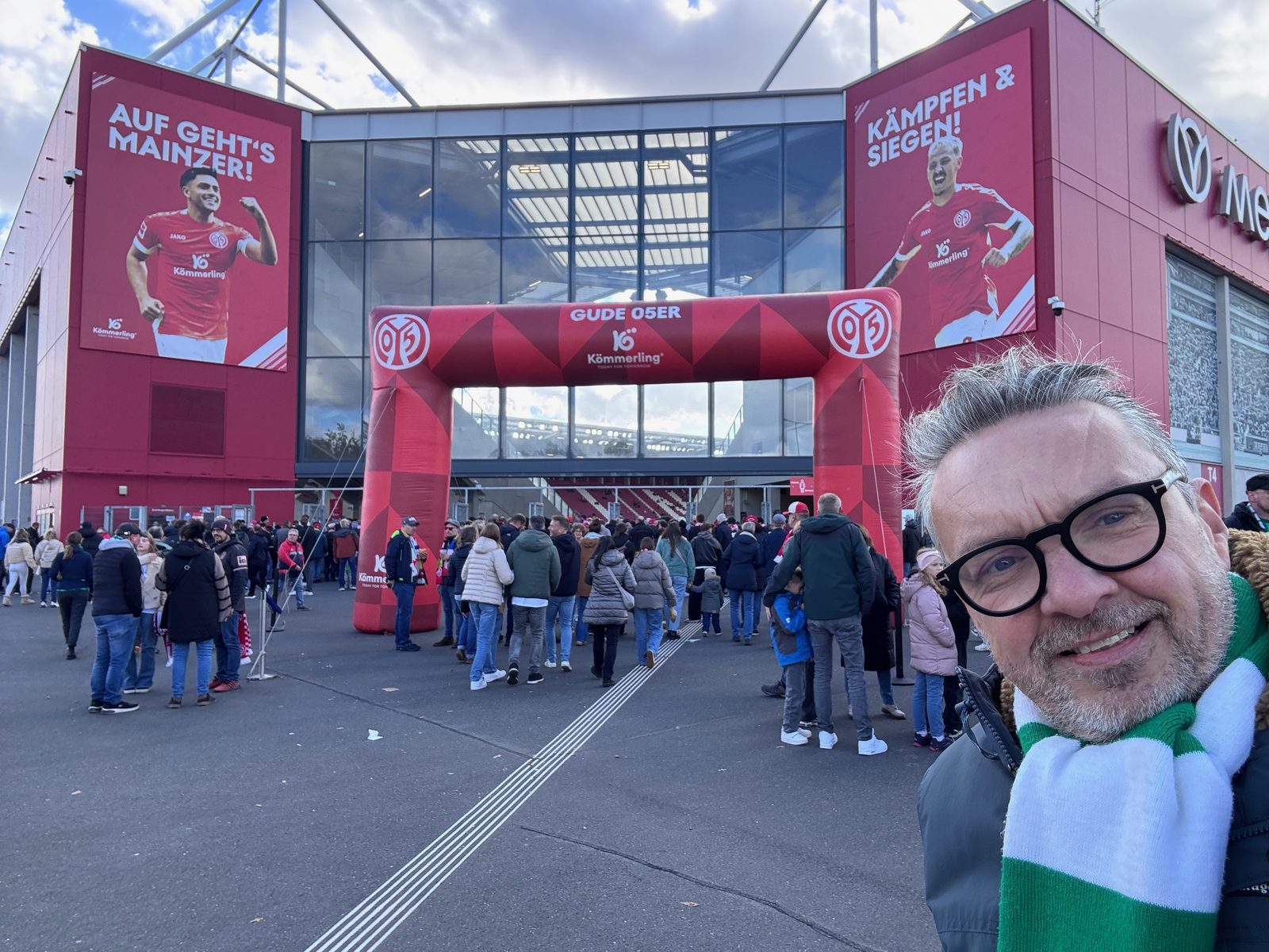 Selfie von Matthias mit grün-weißem Werder-Schal vor dem Eingang der MEWA Arena: ein aufblasbarer roter Torbogen mit „Gude 05er“, an der Fassade große Spielerplakate mit „Auf geht’s Mainzer!“ und „Kämpfen & Siegen!“, davor Zuschauer auf dem Weg ins Stadion.