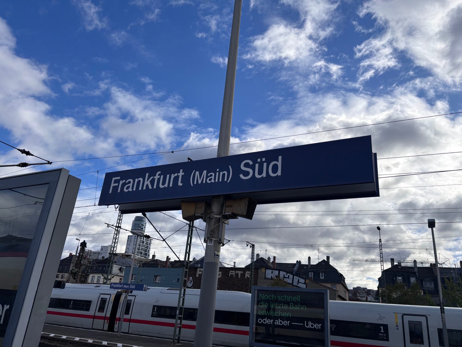 Blaues Bahnsteigschild „Frankfurt (Main) Süd“ vor blauem Himmel mit Wolken, darunter Oberleitungen, ein weißer ICE und Altbauten, im Hintergrund ein Hochhaus.