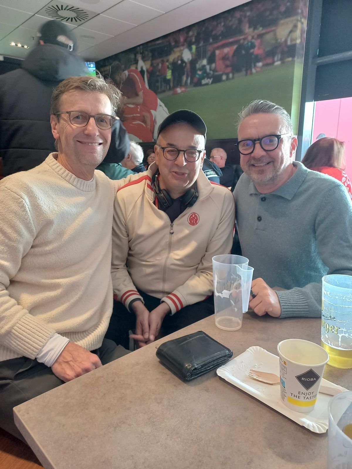 Drei Männer an einem Tisch in einer Stadiongaststätte, alle mit Brille: links Jan im hellen Strickpullover, in der Mitte Daniel Meuren mit Schirmmütze und heller Mainz-05-Trainingsjacke, rechts Matthias im grauen Poloshirt. Auf dem Tisch Becher, ein Pappteller mit Gabel und ein Portemonnaie, an der Wand ein großes Stadionfoto.
