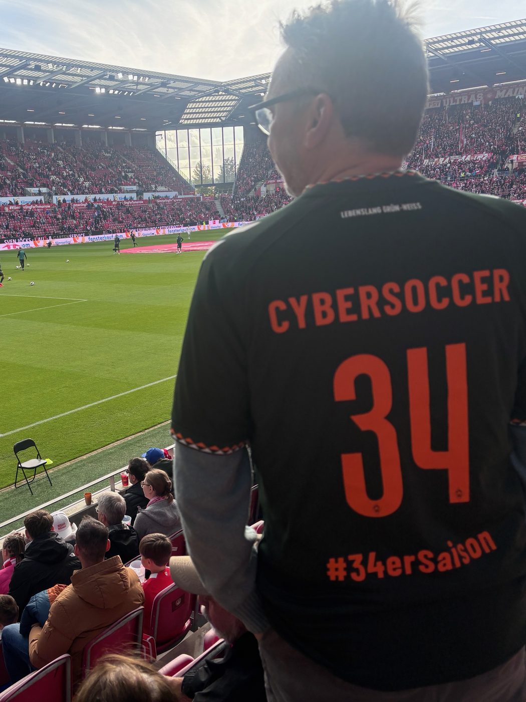 Rückenansicht von Matthias im dunklen Werder-Trikot mit der orangefarbenen Aufschrift CYBERSOCCER, der Nummer 34 und #34erSaison. Er steht auf der Tribüne der MEWA Arena, links unten der Rasen, auf dem sich die Spieler warmlaufen, dahinter die rot besetzten Ränge im Gegenlicht.