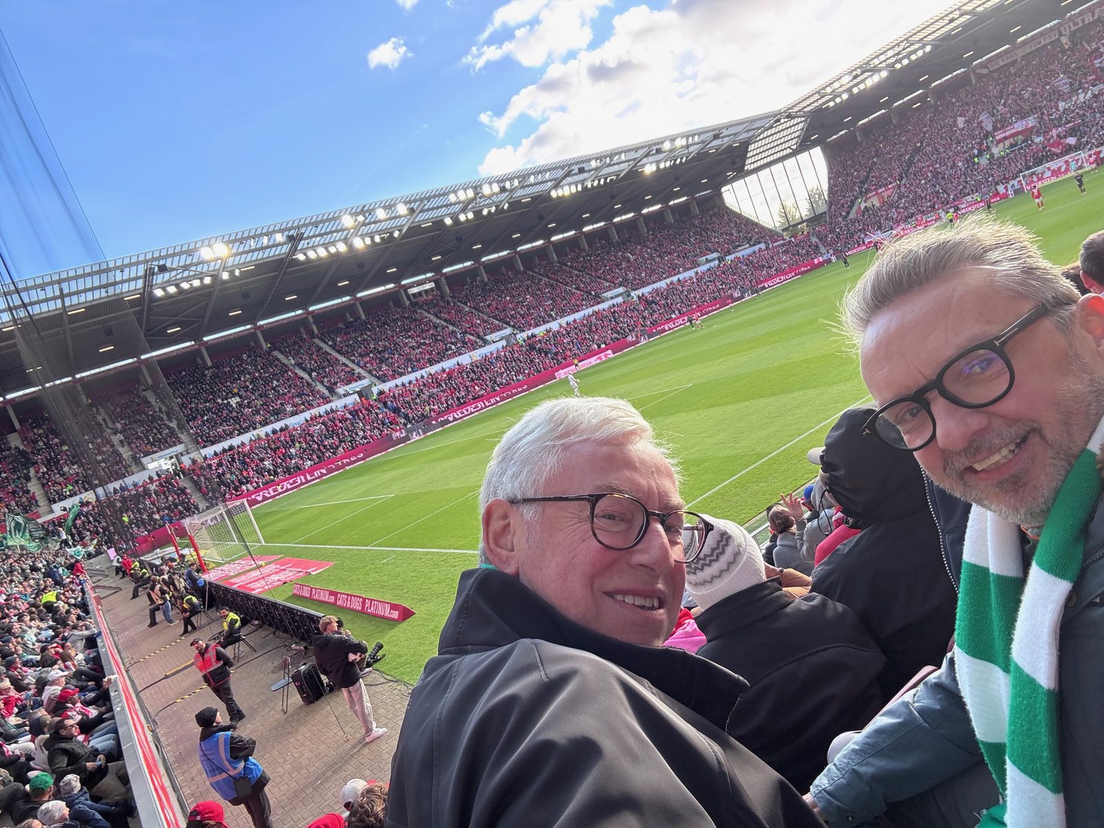 Selfie von Michael Amsinck mit weißem Haar und Brille und Matthias mit grün-weißem Werder-Schal auf der Tribüne der MEWA Arena, hinter ihnen Rasen und die voll besetzte Gegentribüne in Rot unter blauem Himmel.