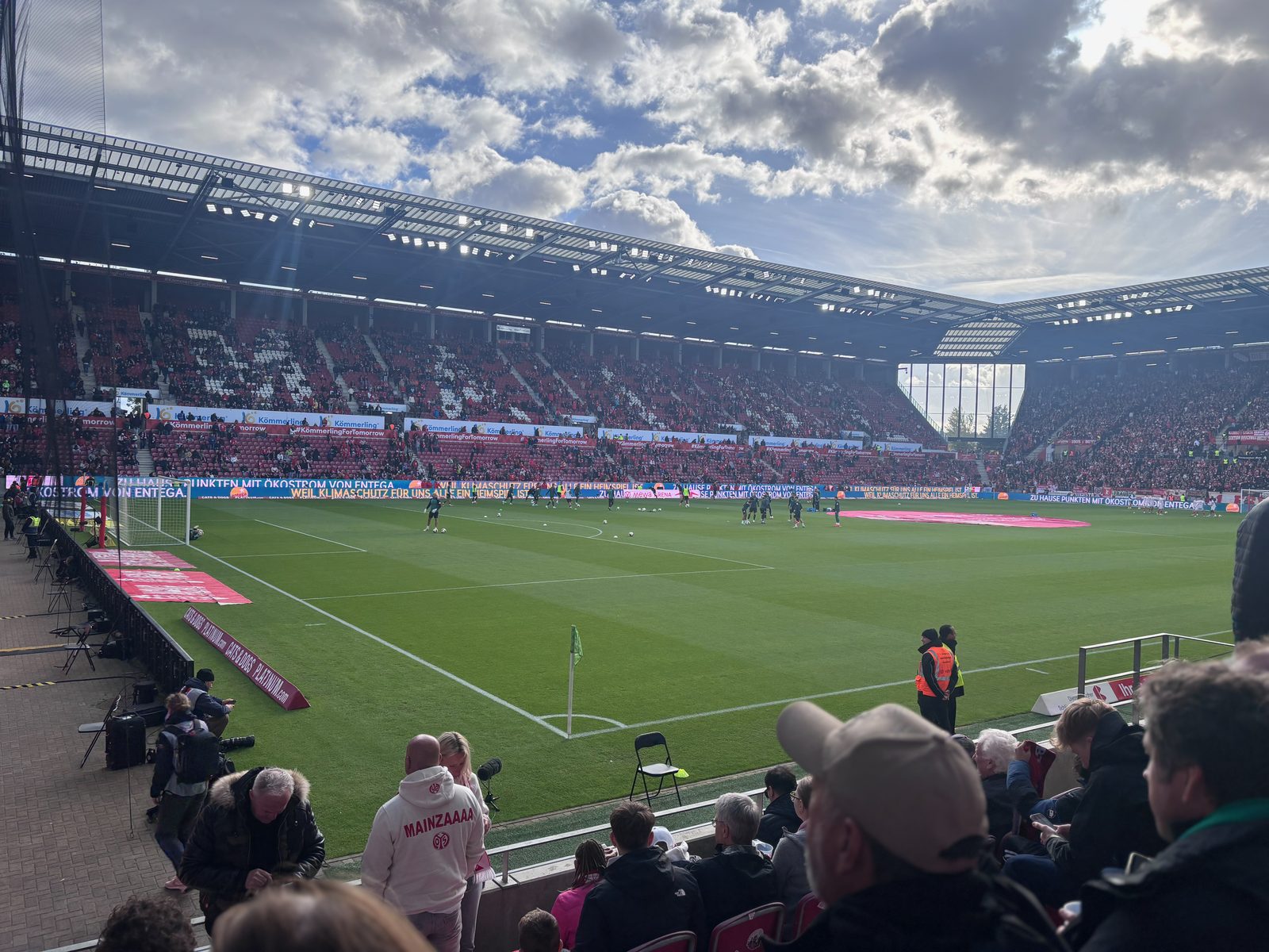 Blick von einem Platz nahe der Eckfahne flach über den Rasen der MEWA Arena, auf dem sich die Spieler warmlaufen, dahinter die Gegentribüne unter wolkigem Himmel, im Vordergrund Zuschauer und Ordner.