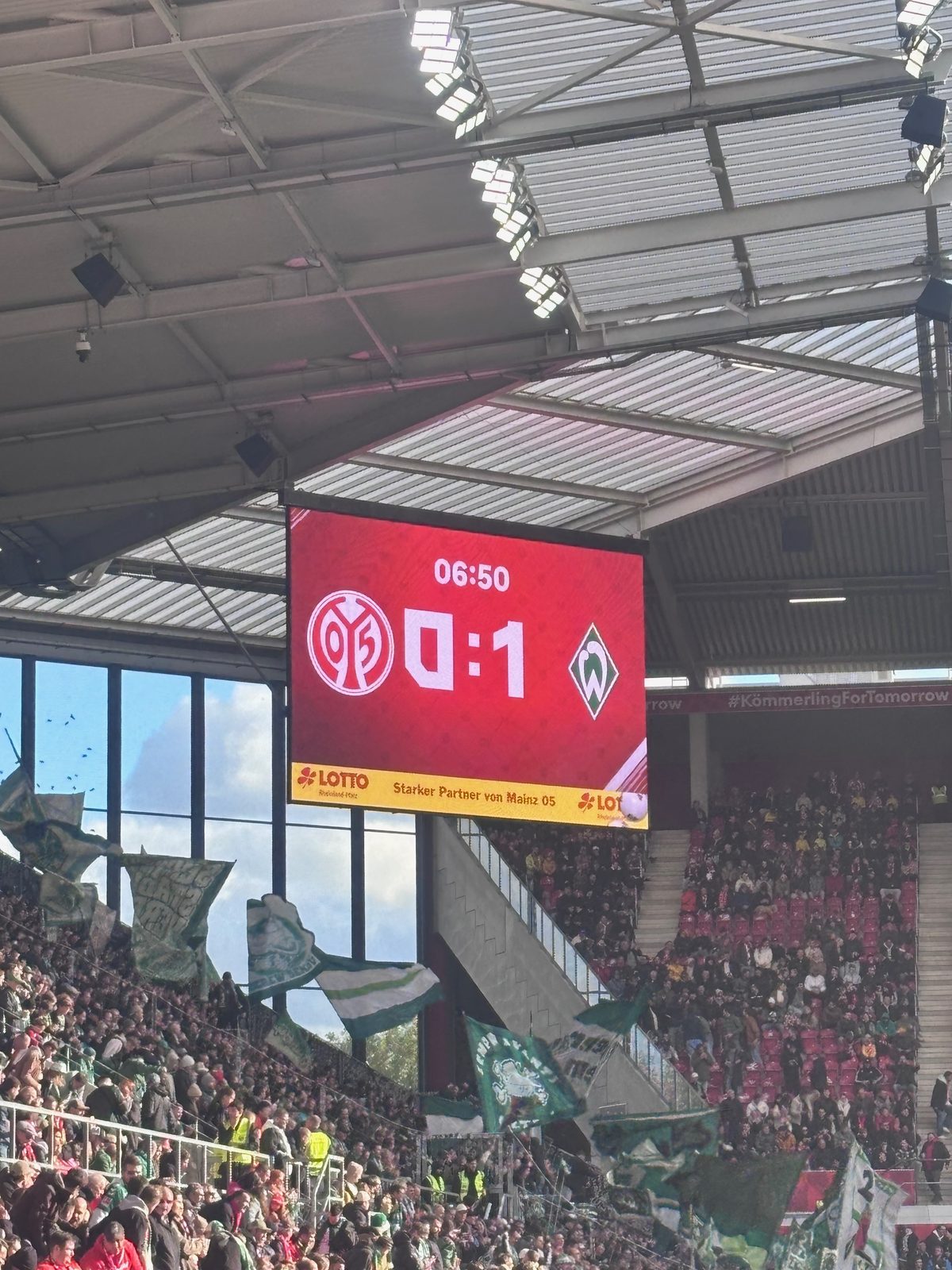 Die Anzeigetafel unter dem Stadiondach zeigt nach 6:50 Minuten 0:1 zwischen dem Mainz-05- und dem Werder-Wappen, darunter der Gästeblock mit grün-weißen Fahnen.