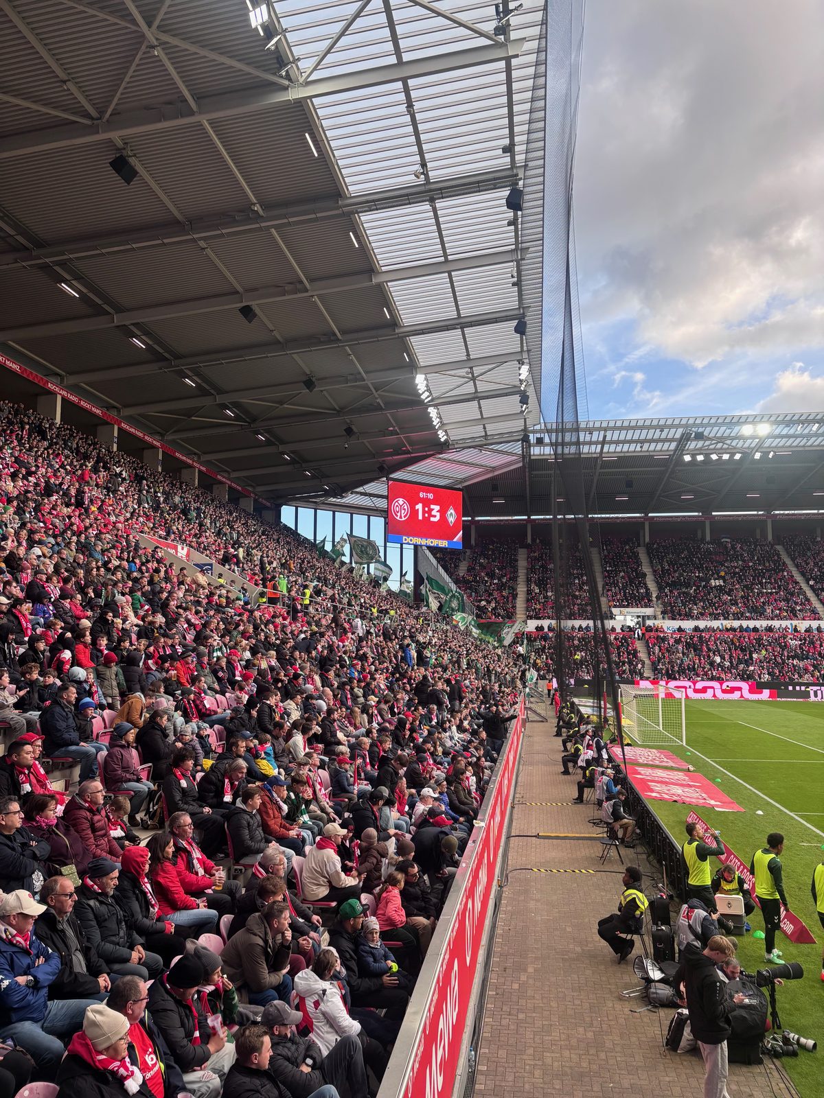 Blick entlang der voll besetzten Haupttribüne zum Stadiondach, die Anzeigetafel zeigt nach 61:10 Minuten 1:3, rechts die Werder-Ersatzspieler in gelben Leibchen am Spielfeldrand.